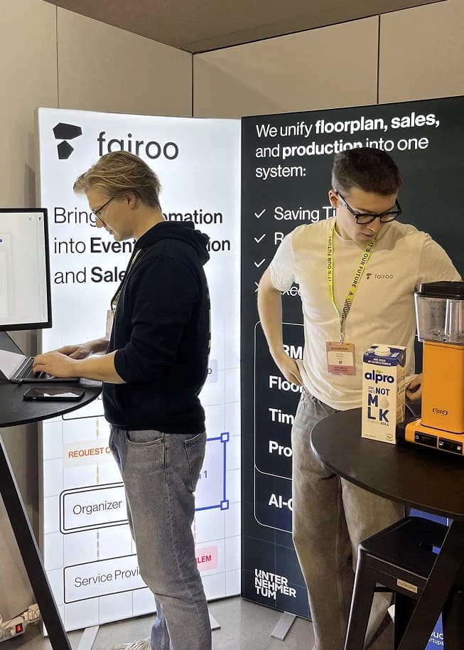 Zwei Teammitglieder am Fairoo-Messestand, einer arbeitet am Laptop vor dem Fairoo-Roll-up